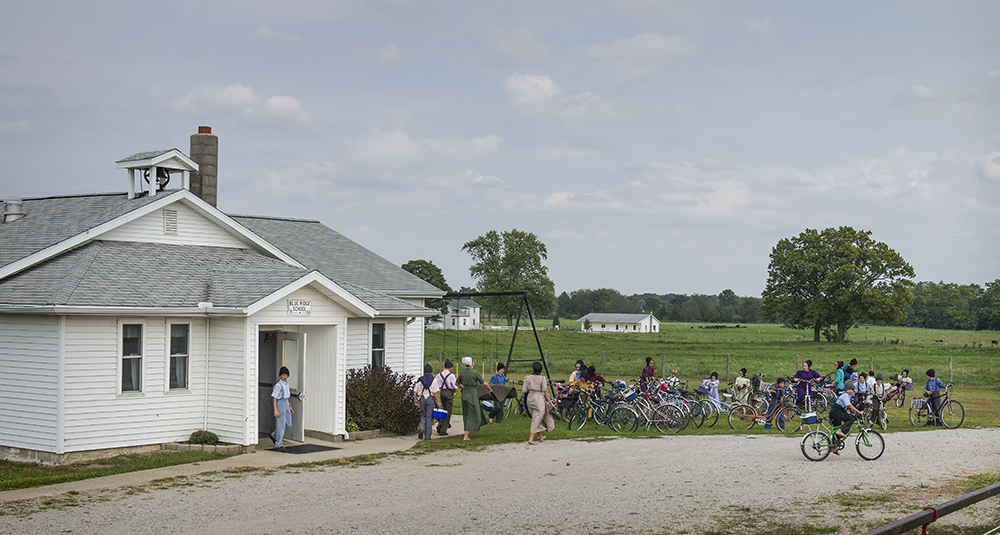 Amish school & yard many children & adults in playground and on bicycles in country